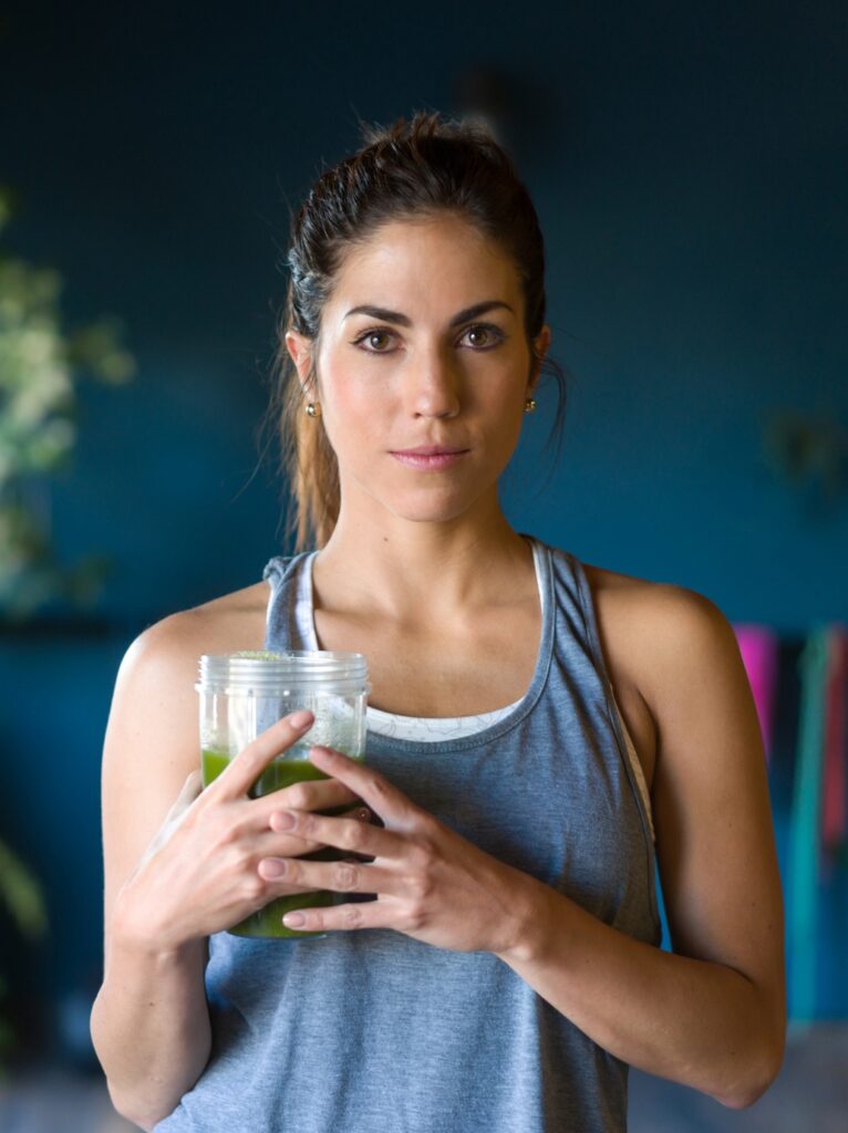 Eine junge Frau, die in einem Fitnessraum steht und einen grünen Smoothie in der Hand hält. Gesunder Lebensstil.